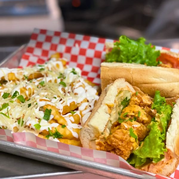 Creole fried shrimp po'boy with lettuce, tomato, and a side of loaded fries on a metal tray.