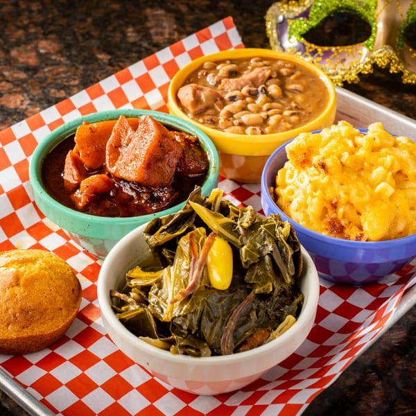 A tray of Creole dishes including candied yams, black-eyed peas, mac and cheese, collard greens, and cornbread.
